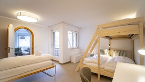 Ein Schlafzimmer mit Etagenbett, Schreibtisch und Stuhl. Ein Fenster und eine Tür zu einem anderen Raum sind vorhanden.
