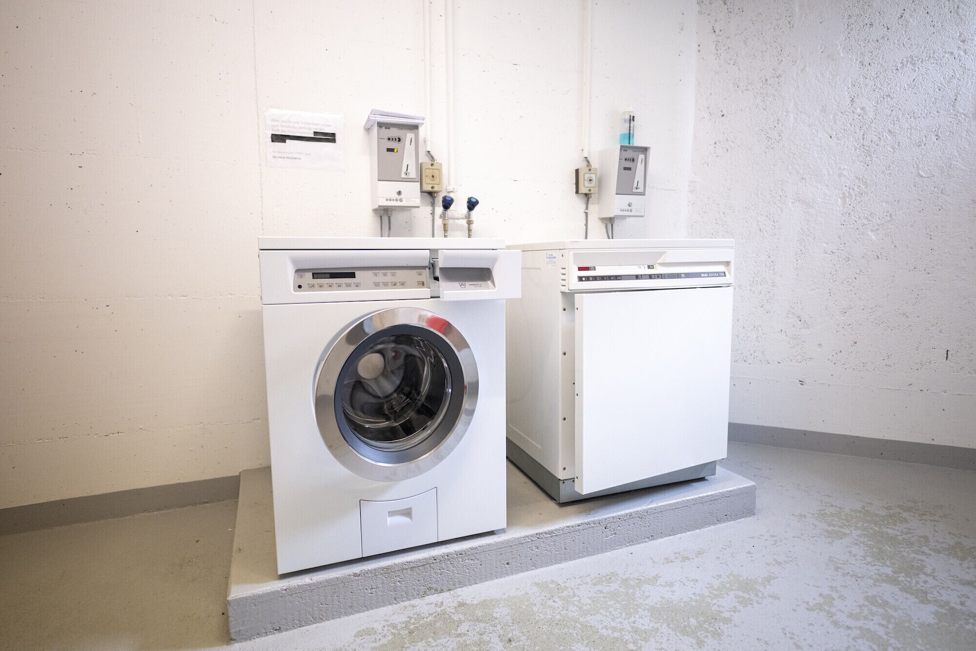 Een wasserij met twee wasmachines en drogers, een bedieningspaneel en een waterverwarmer.