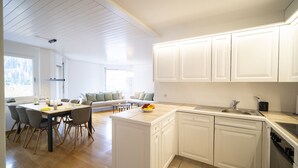 Eine moderne Küche mit weißen Schränken, ein Essbereich mit Holztisch und -stühlen sowie ein Blick auf eine grüne Außenanlage durch ein großes Fenster.