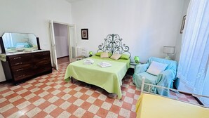 Une chambre d’hôtel avec un sol à carreaux, un lit avec une literie verte, une commode en bois, une chaise et une petite table avec une lampe.