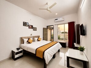 A hotel room with a bed, bedside table, a desk, a ceiling fan, an air conditioning unit, a window with curtains, and wall-mounted artwork. - Hotel Arul's (Udaipur)
