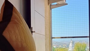 Una cama con ropa de cama blanca y una almohada, una ventana con vista a un edificio y árboles, y un pasamanos de metal.