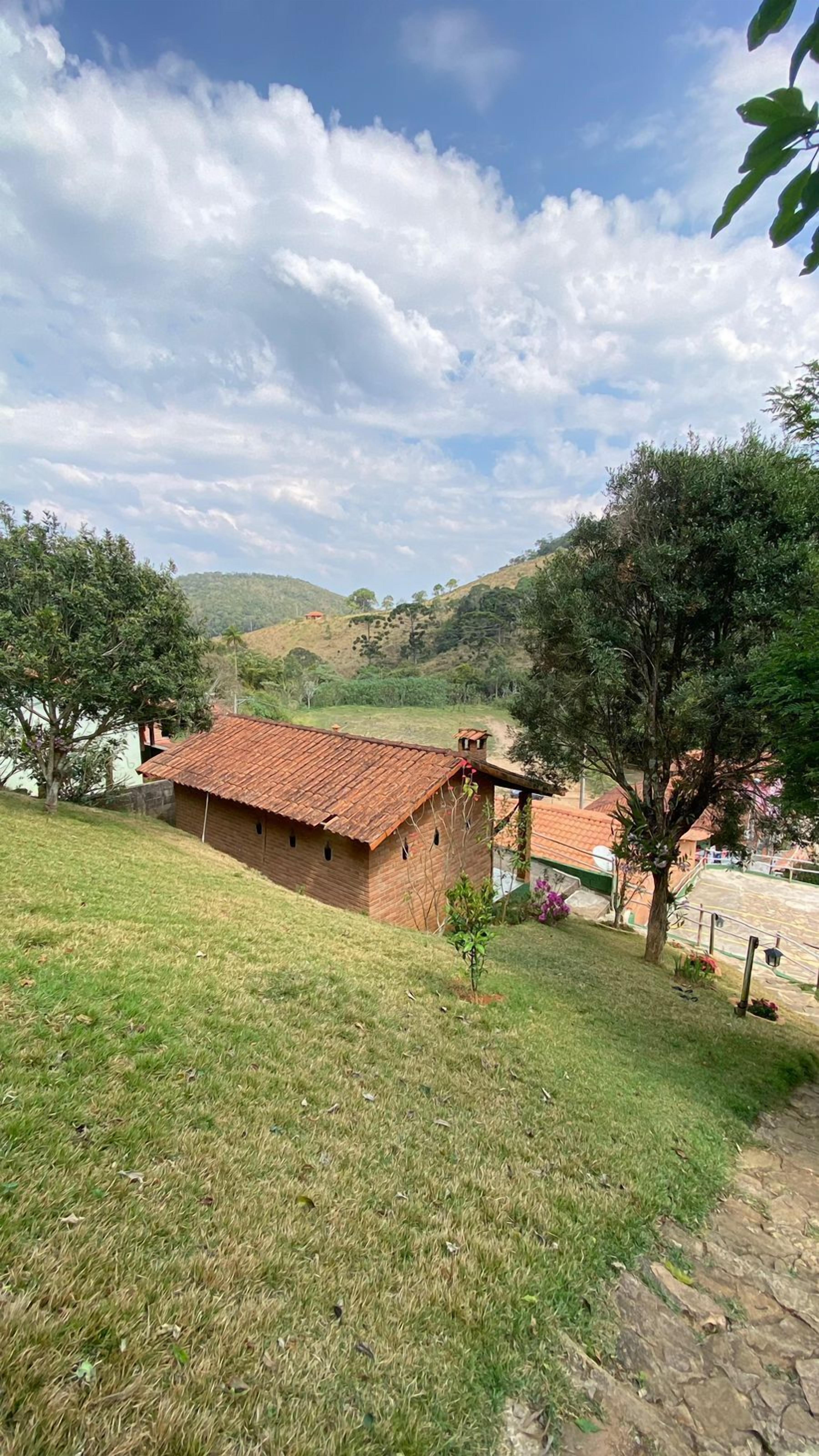 A rustic house with a tiled roof, surrounded by a grassy area and trees, set against a backdrop of hills and a partly cloudy sky.
