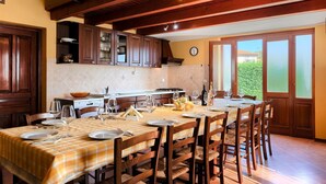 Een eetgedeelte met een lange tafel voor zes personen, houten stoelen, een keuken op de achtergrond en uitzicht op groen door een glazen deur.