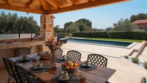 Ein Essbereich im Freien mit Blick auf einen Pool, mit einem Holztisch, der mit Tellern, Tassen und einer Obstschale gedeckt ist.