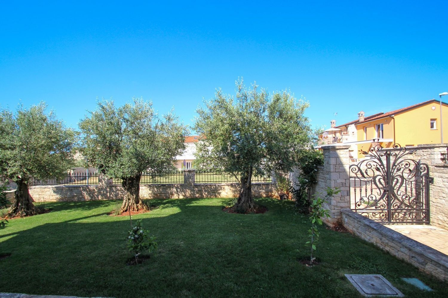 Un jardin bien entretenu, avec un mur de pierre, un portail en métal et, au fond, un bâtiment à la façade jaune.