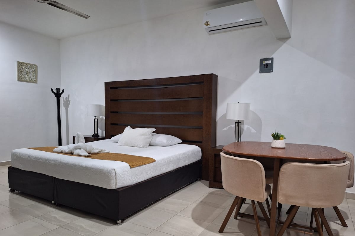 A hotel room with a bed, bedside tables, a lamp, a small table with chairs, and an air conditioning unit.