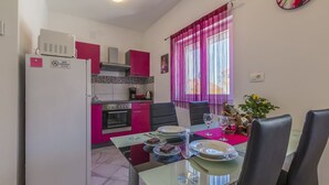 A compact kitchen with pink cabinets, a dining table set for two, and a window with purple curtains. - Charming dog-friendly flat with private balcony, playground, barbecue (Crikvenica)