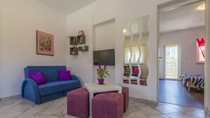 A modern living room with a blue sofa, purple cushions, a white coffee table, and a potted plant. There is a TV mounted on the wall, a small shelf with decorative items, and a mirror with a wavy frame. - Charming dog-friendly flat with private balcony, playground, barbecue (Crikvenica)