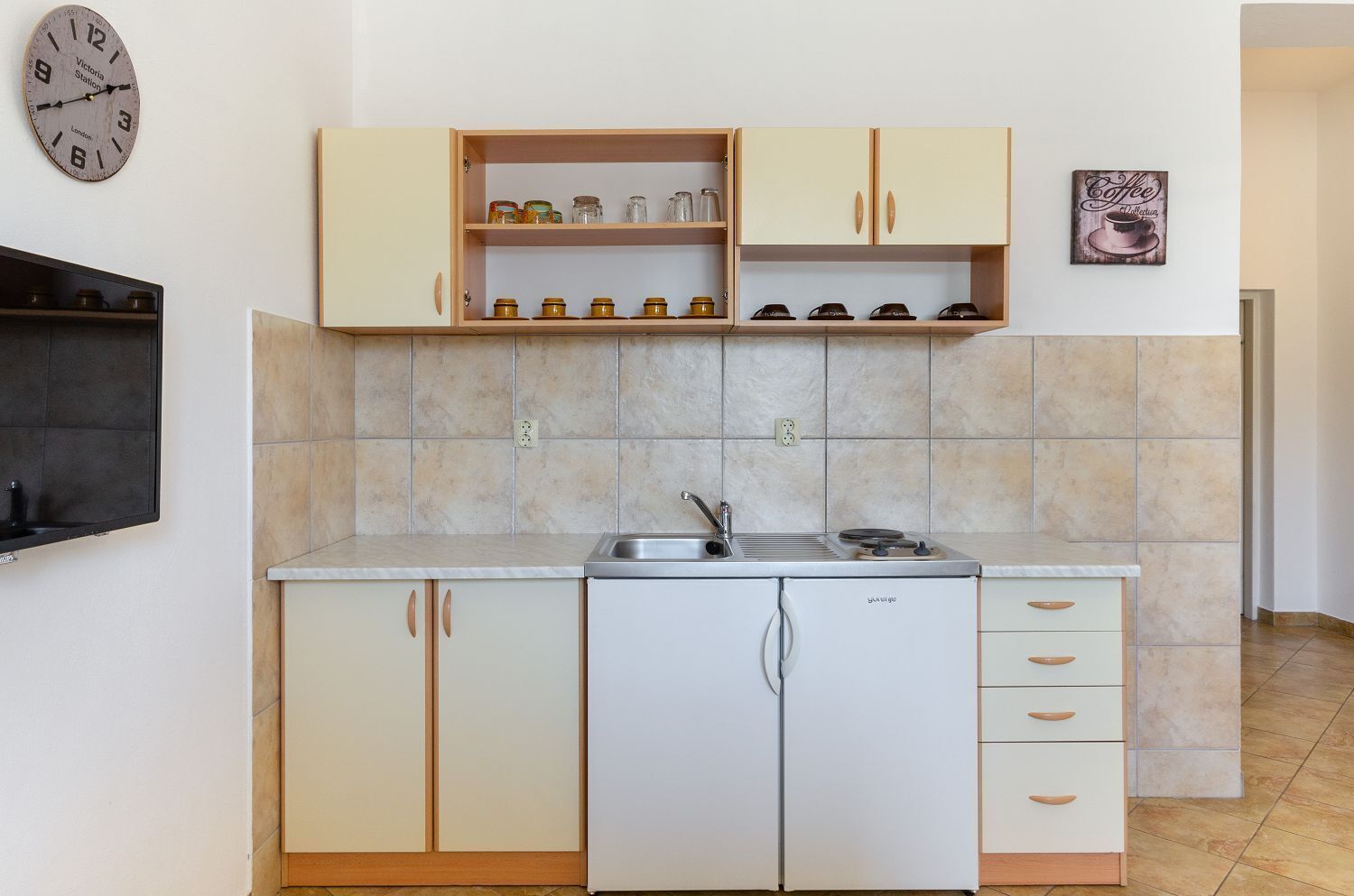 Eine kompakte Küche mit weißen Schränken, einem eingebauten Backofen und einem Spülbecken. An der Wand hängt eine Uhr und über den Schränken ein gerahmtes Bild.