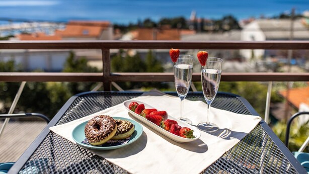 A table with a plate of strawberries and chocolate donuts, two glasses of water with strawberries, and a scenic view of the sea and buildings.