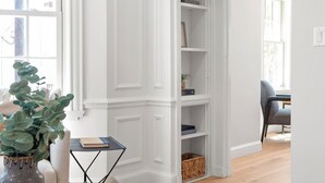 Ein Zimmer mit Holzboden, weißen Wänden, einem Bücherregal, einem kleinen Tisch mit Vase und Büchern sowie einem Stuhl.