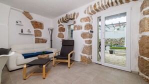 Sala de estar com sofá branco, uma cadeira preta e uma pequena mesa. As paredes são de pedra e há uma porta de vidro que dá para uma área externa.