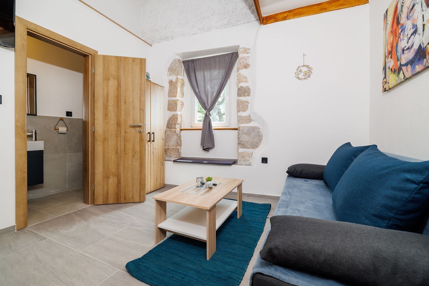 A cozy living room with a wooden door, a stone wall, a window with curtains, a blue sofa, and a small table.