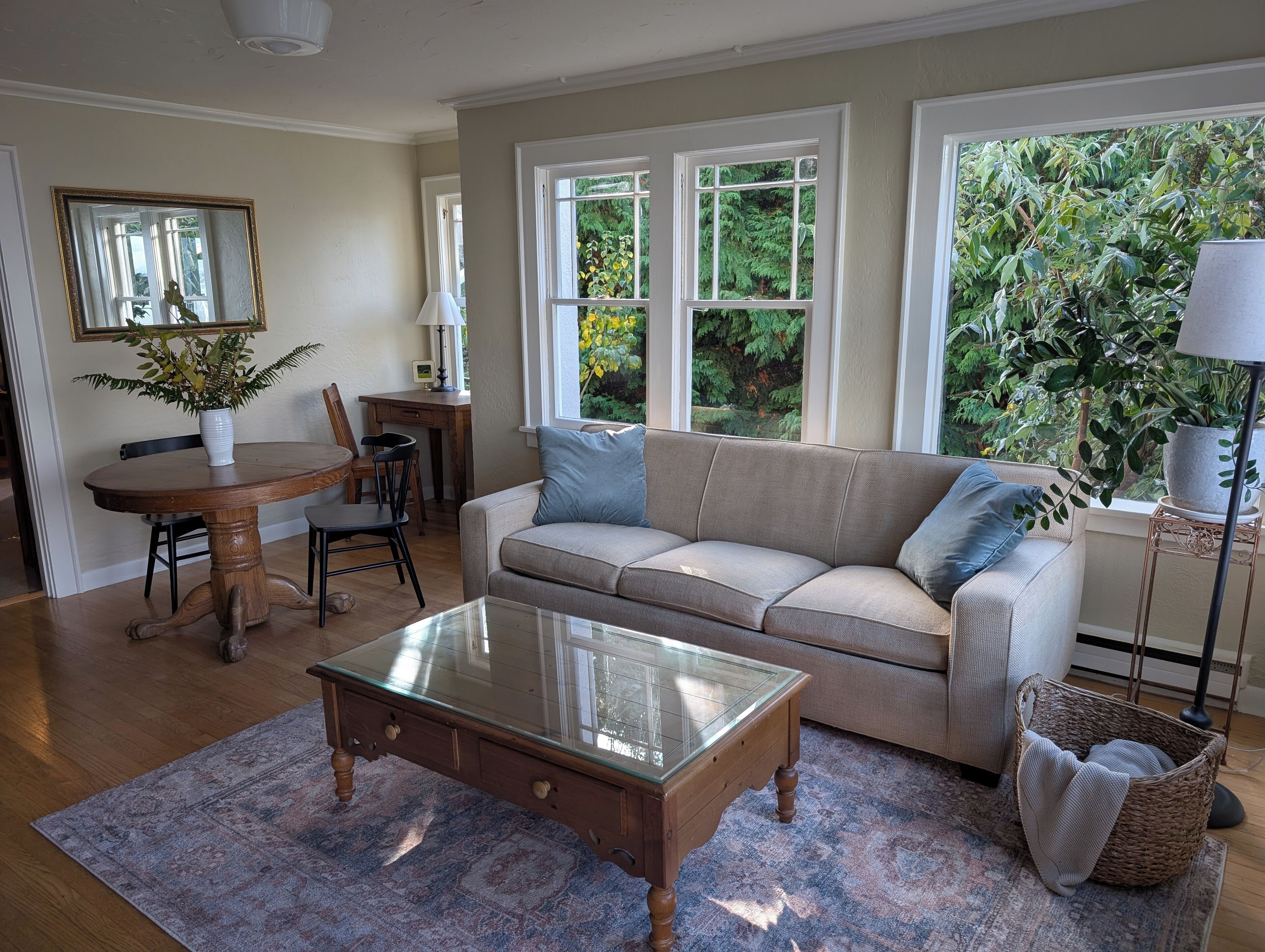 A living room with a beige sofa, a glass coffee table, a dining table with chairs, and a view of greenery through large windows.
