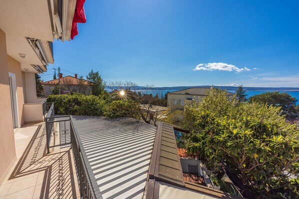 A rooftop terrace with a view of the sea, surrounded by greenery and buildings.