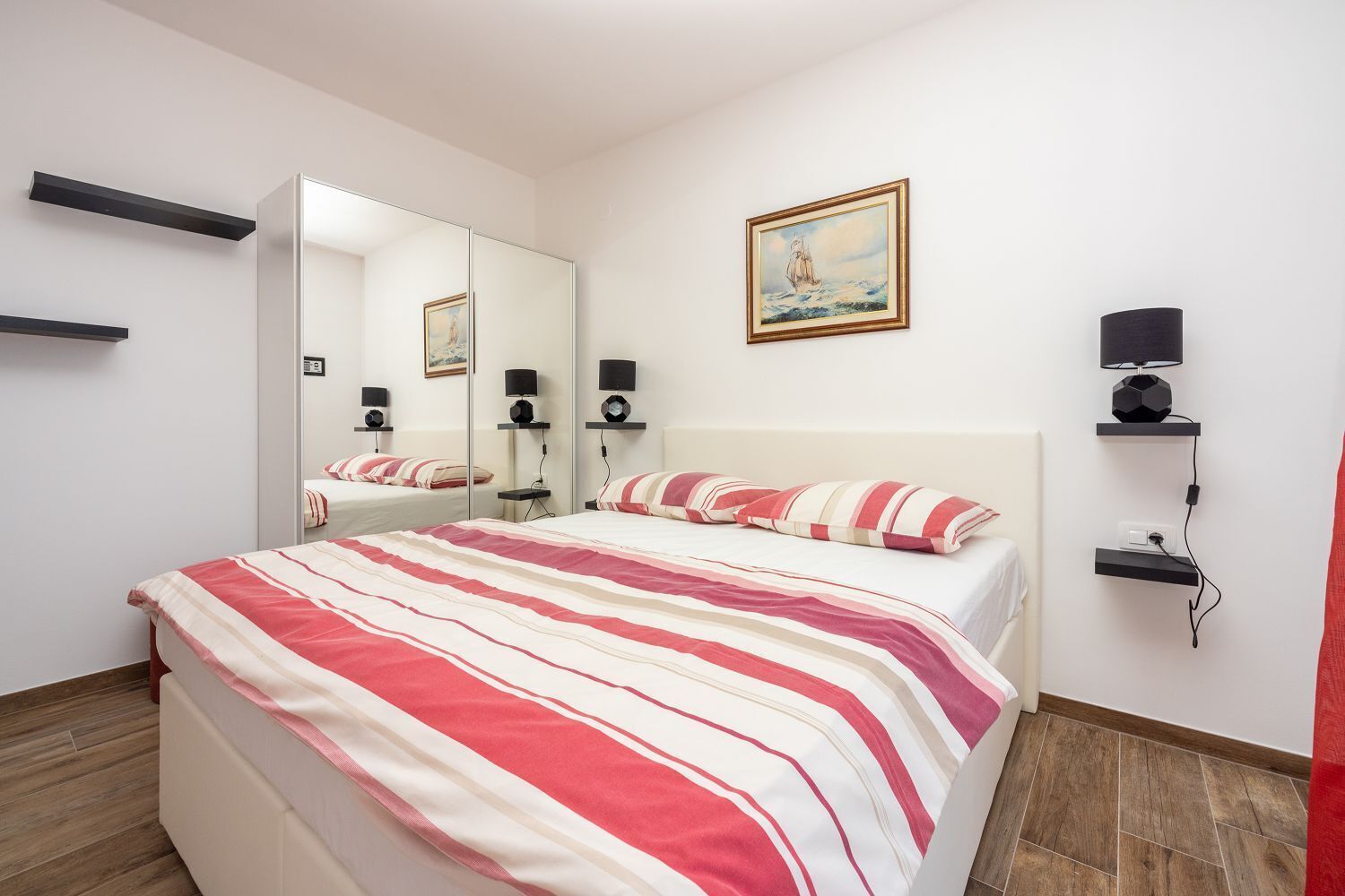 Habitación de hotel con una cama grande, una mesita de noche, un espejo, estante con lámpara y un cuadro.