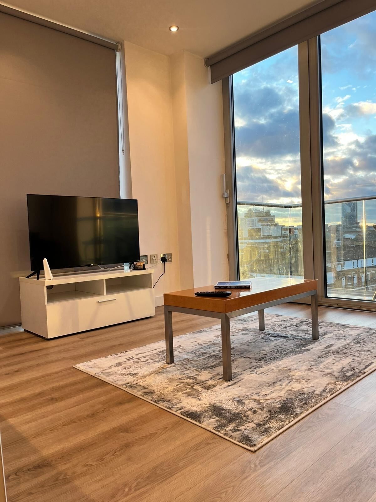 Un saló modern amb un televisor de pantalla plana sobre un moble blanc, una taula baixa de fusta i una finestra gran que ofereix vista a la ciutat.