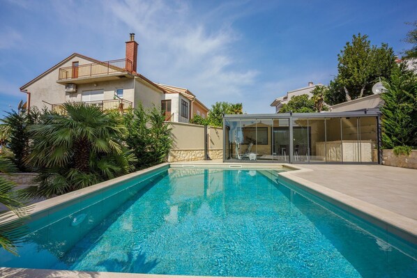 A swimming pool with a glass enclosure, a house with a balcony, and surrounding greenery.