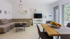 Ein modernes Wohnzimmer mit einem Ecksofa, einem runden Couchtisch, einem Fernseher auf einem weißen Lowboard und einem Esstisch mit Obstschale.