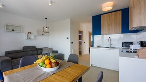 A modern living room with a grey sofa, a dining table with a fruit bowl, and a kitchen with white cabinets and a blue accent wall. - Impressive flat just 500 metres from the beach with sea views, sauna, gym, pool (Crikvenica)