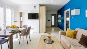 A modern living room with a blue accent wall, a beige sofa with a yellow pillow, a dining area with a fruit bowl, and a kitchen with a TV and wooden cabinets. - Exquisite flat with sea view, sauna, gym, pool, air-conditioned bedrooms - only 500 metres to the beach (Crikvenica)