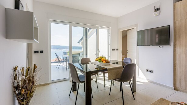 A modern dining area with a glass table, chairs, and a bowl of fruit. There is a TV mounted on the wall and a view of the outdoors through large windows. - Exquisite flat with sea view, sauna, gym, pool, air-conditioned bedrooms - only 500 metres to the beach (Crikvenica)