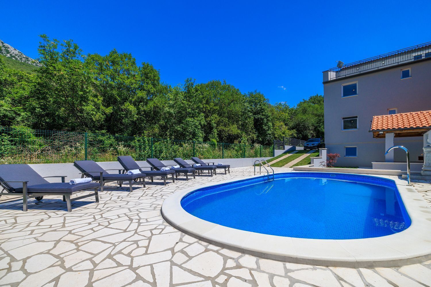 A swimming pool with lounge chairs, a stone-tiled deck, and a building with a red-tiled roof in the background.