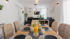 A modern dining area with a wooden table set for four, surrounded by beige chairs. A bottle of orange juice and a bowl of fruit are on the table.