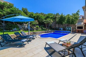Un patio trasero con piscina, sillas de descanso, zona de barbacoa y un muro de piedra.