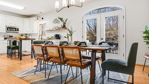 Et moderne køkken med hvide skabe, et spiseområde med træstole, et stort vindue med udsigt og en lysekrone.
