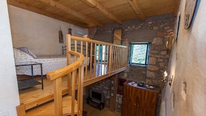 Una habitación rústica con una escalera de madera, una litera, una pared de piedra y un ventanal pequeño.
