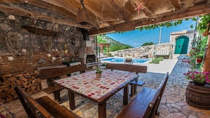Un patio exterior con suelo de piedra, bancos de madera, una mesa con mantel estampado de frutas, una pared de piedra con horno de ladrillo y vistas a una piscina y edificios de piedra al fondo.