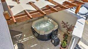 Un jacuzzi bajo una pérgola de madera con suelo de piedra, rodeado de plantas en macetas y una silla.