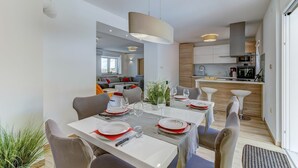 Un comedor moderno con una mesa rectangular dispuesta para cuatro personas, rodeada de sillas. Se ve una cocina al fondo con armarios de madera y un horno empotrado.