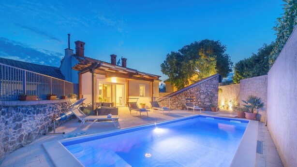 Una piscina con sillas de descanso, un muro de piedra y un edificio con fachada de piedra y techo de tejas.