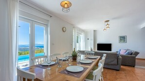 Comedor con mesa de madera dispuesta para cuatro personas, sillas blancas y vistas al mar a través de amplios ventanales.