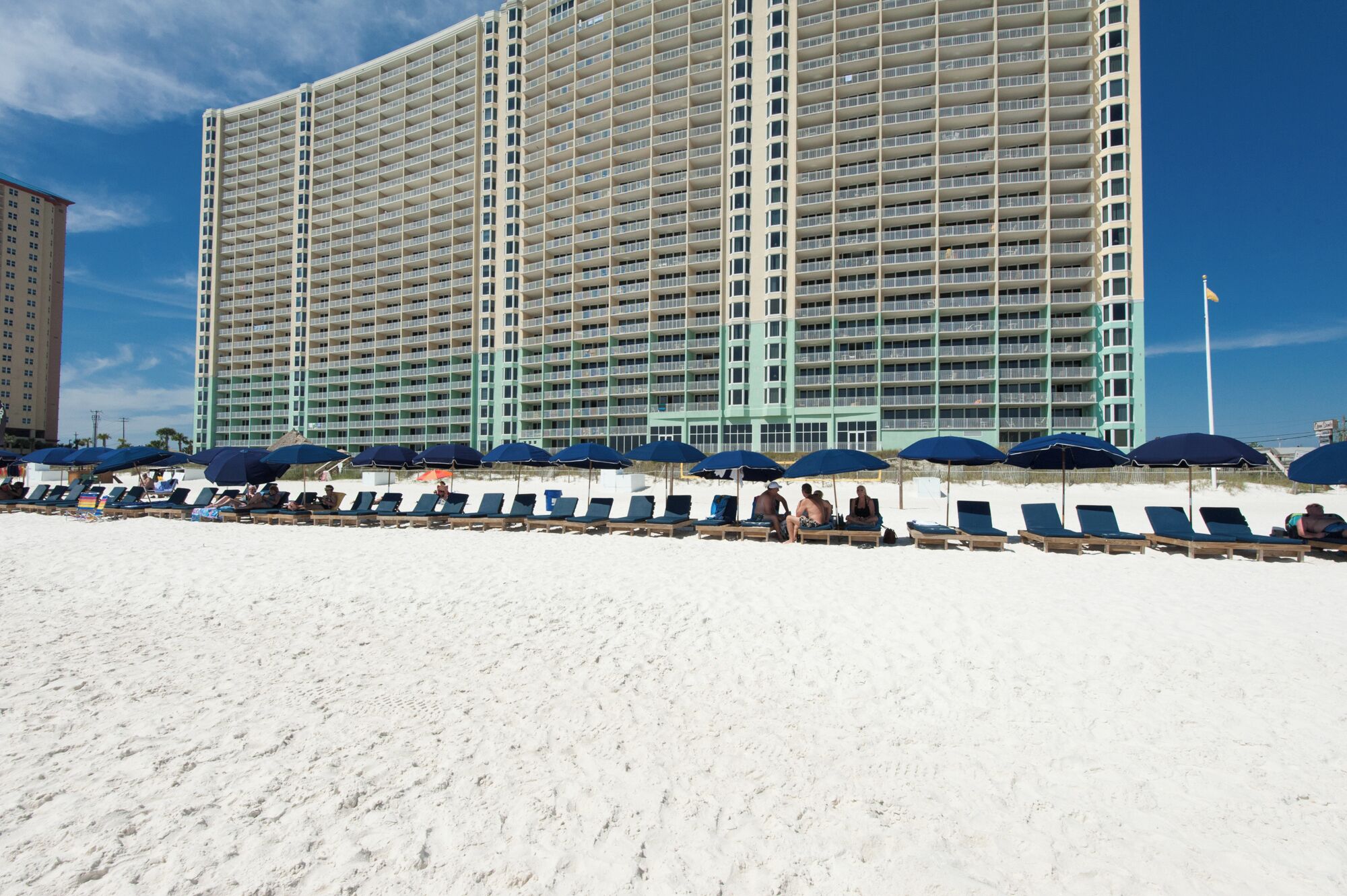 A large hotel with multiple floors and balconies, a sandy beach in the foreground with lounge chairs and umbrellas, and a clear blue sky.