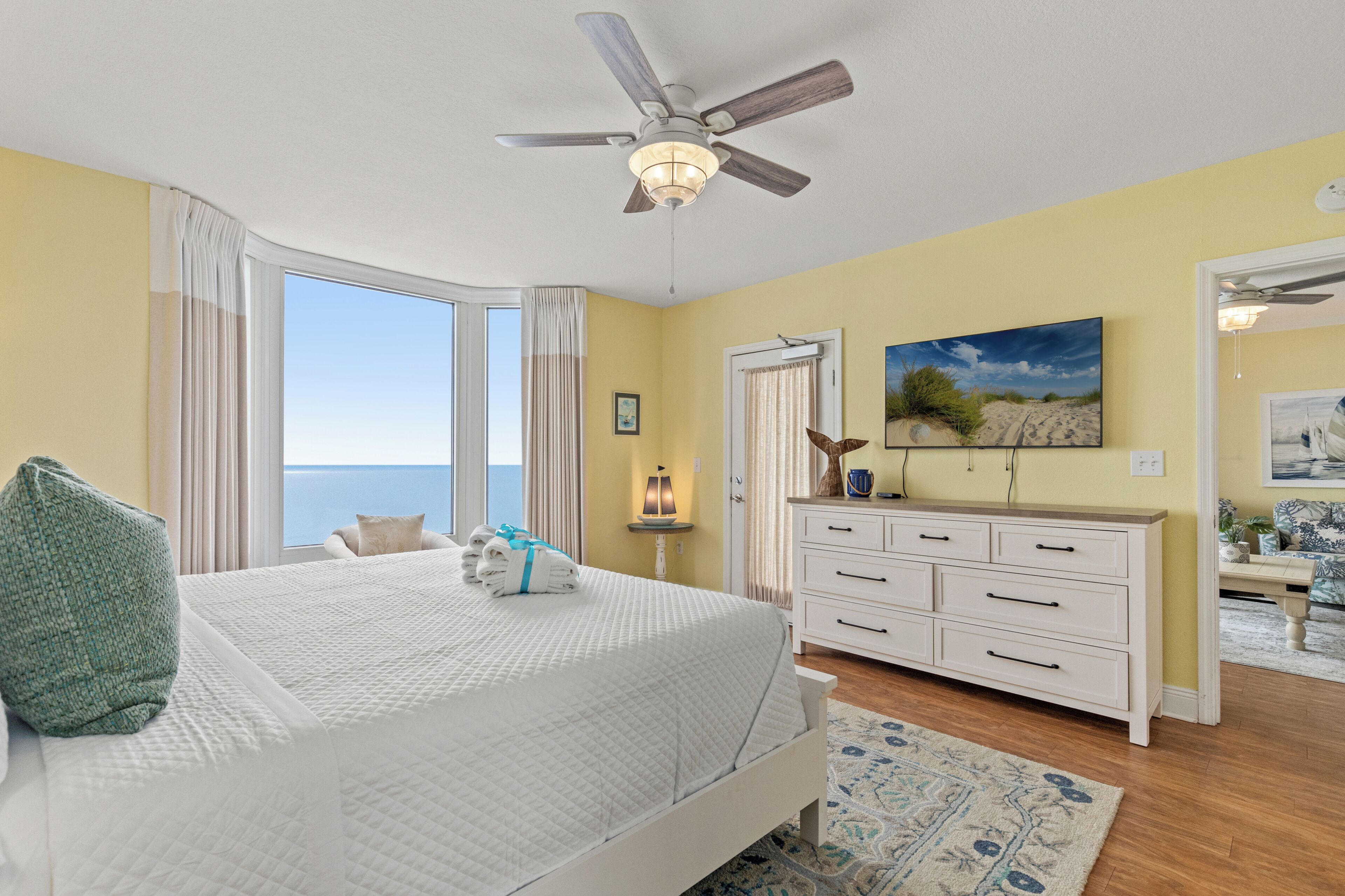 A bedroom with a bed, a dresser, a ceiling fan, a window with curtains, and a painting on the wall.