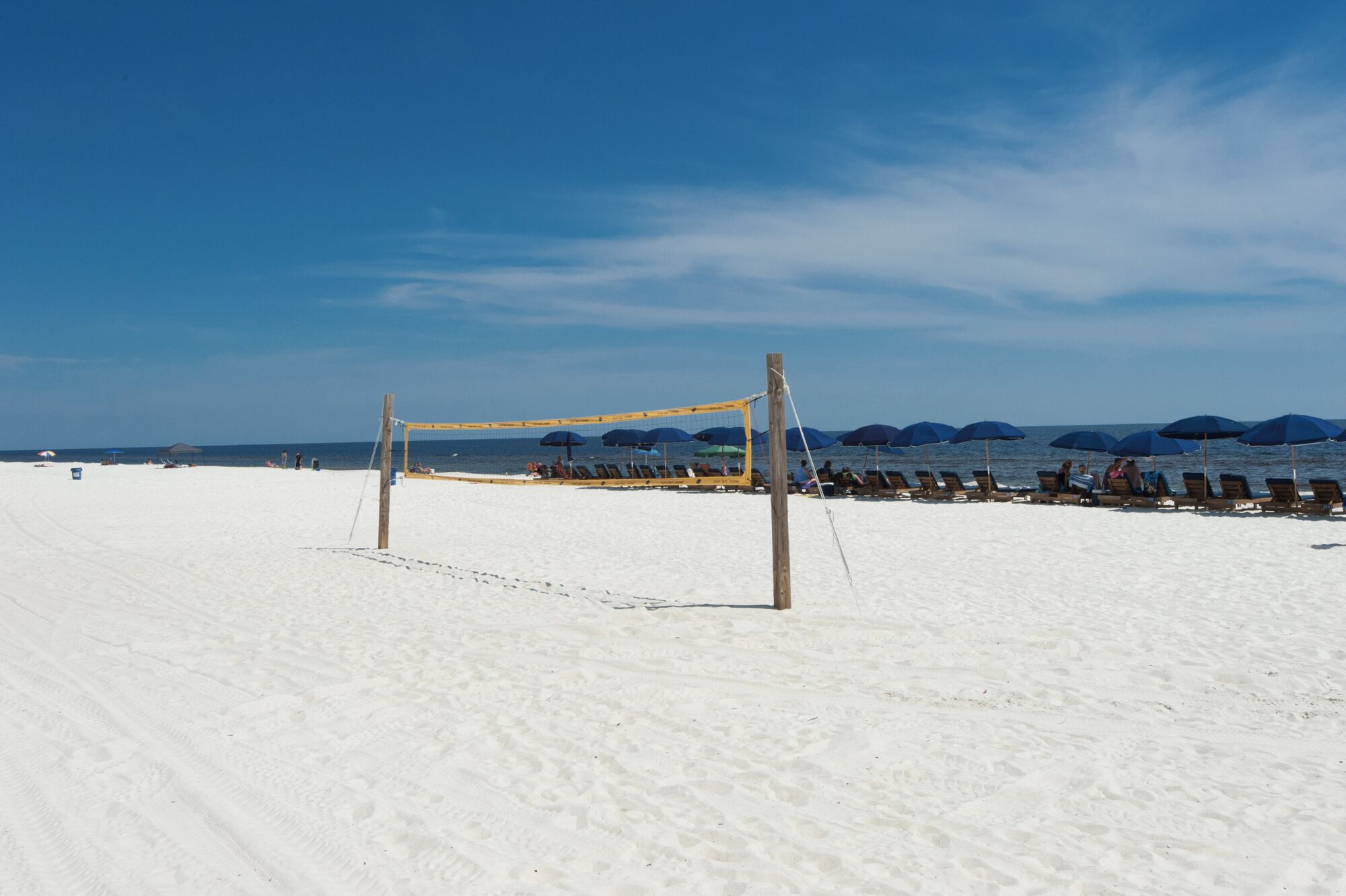 Uma rede de voleibol de praia numa praia arenosa, com guarda-sóis e espreguiçadeiras ao fundo.
