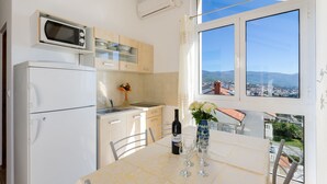 Una cucina con frigorifero, microonde e pensili, un tavolo da pranzo con una bottiglia di vino e bicchieri, e vista su una città attraverso una finestra.