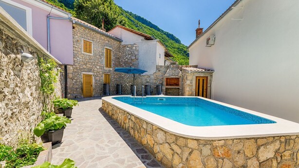 Une piscine en pierre, entourée de murs de pierre et de plantes en pot, offrant une vue sur un paysage montagneux.