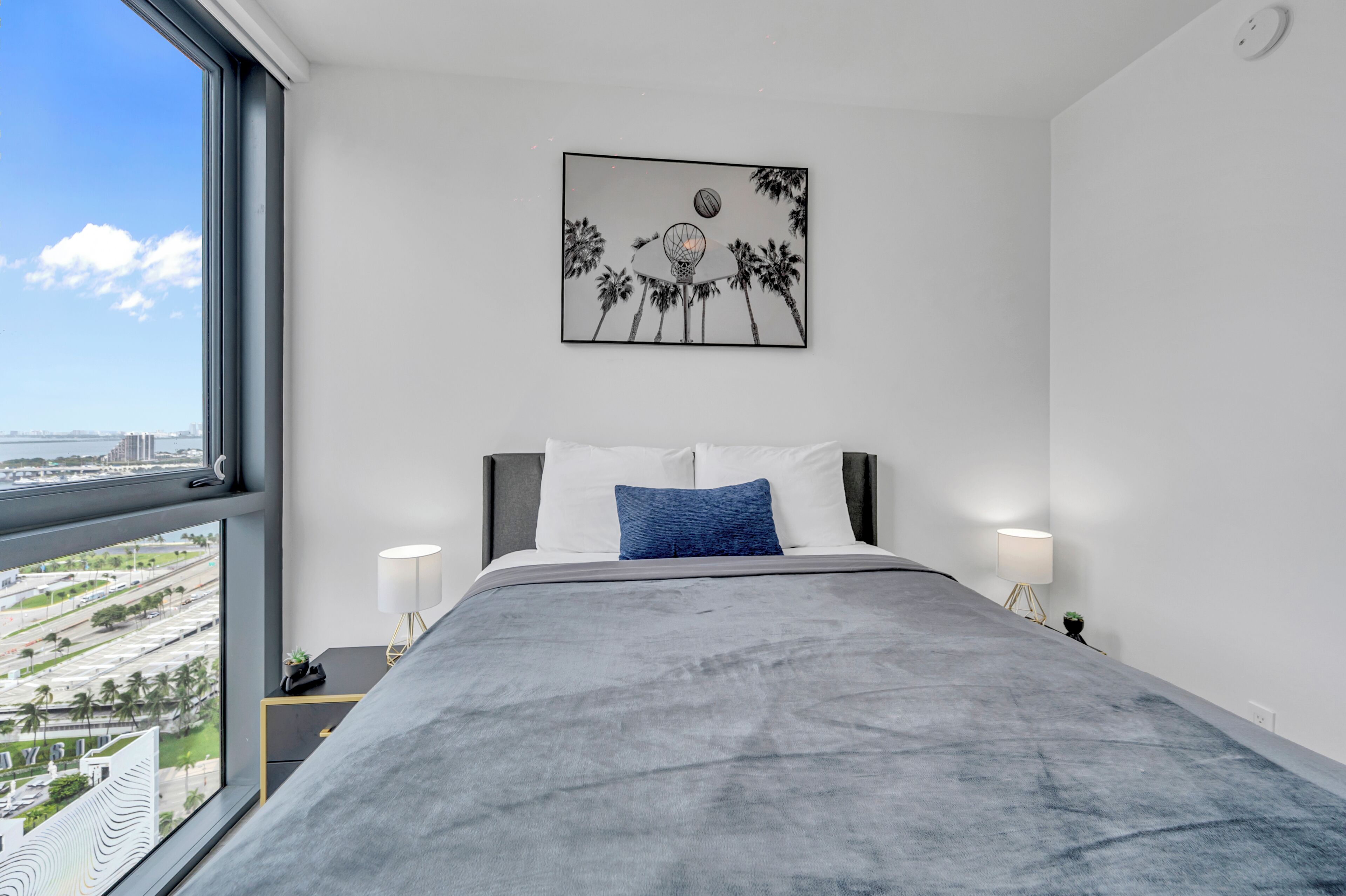 A modern bedroom with a large window, a bed with grey bedding, a blue pillow, and a wall-mounted artwork depicting palm trees and a basketball hoop.