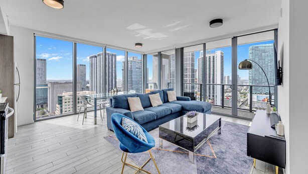 Ein modernes Wohnzimmer mit großem Fenster, einem blauen Sofa, einem Glastisch und Blick auf hohe Gebäude.