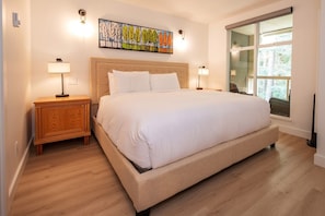 A bedroom with a large bed, wooden nightstands, a wall-mounted painting, and a window with a view.