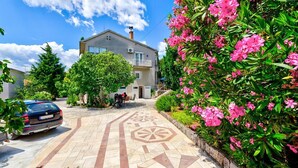 Una calle residencial con un coche estacionado, una casa con balcón y un jardín con flores rosas.