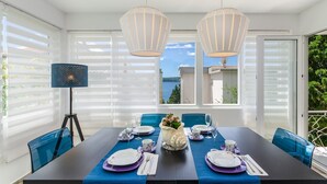 Un comedor con una mesa dispuesta para cuatro, ventanas amplias con persianas y vista al mar.