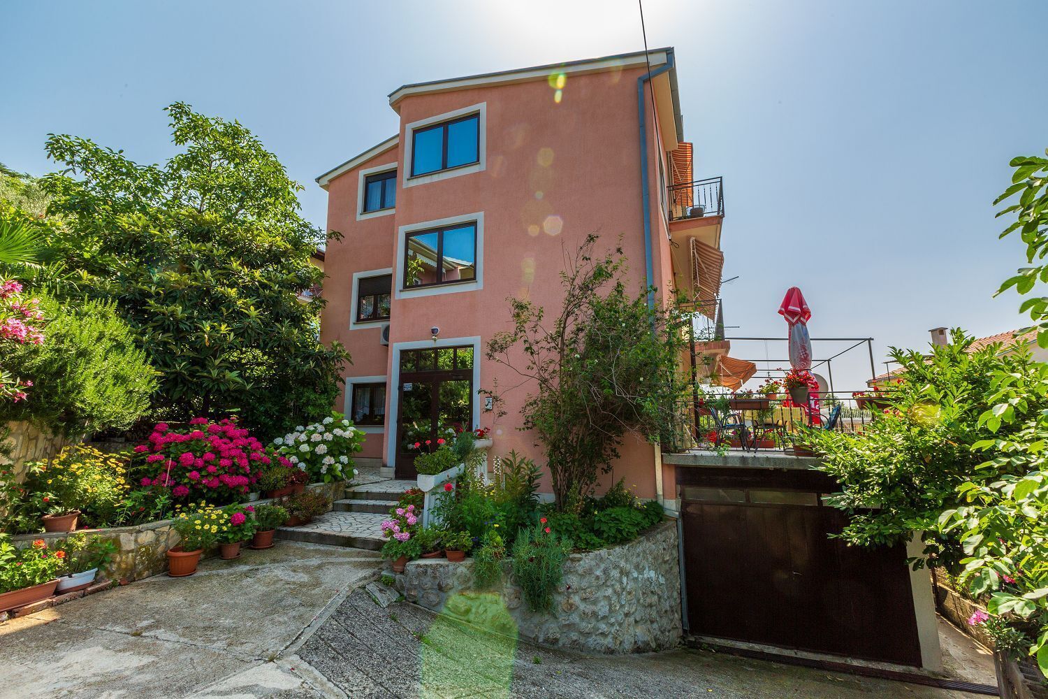A multi-story building with a pink facade, a balcony with a red umbrella, and a garden with various potted plants and flowers.