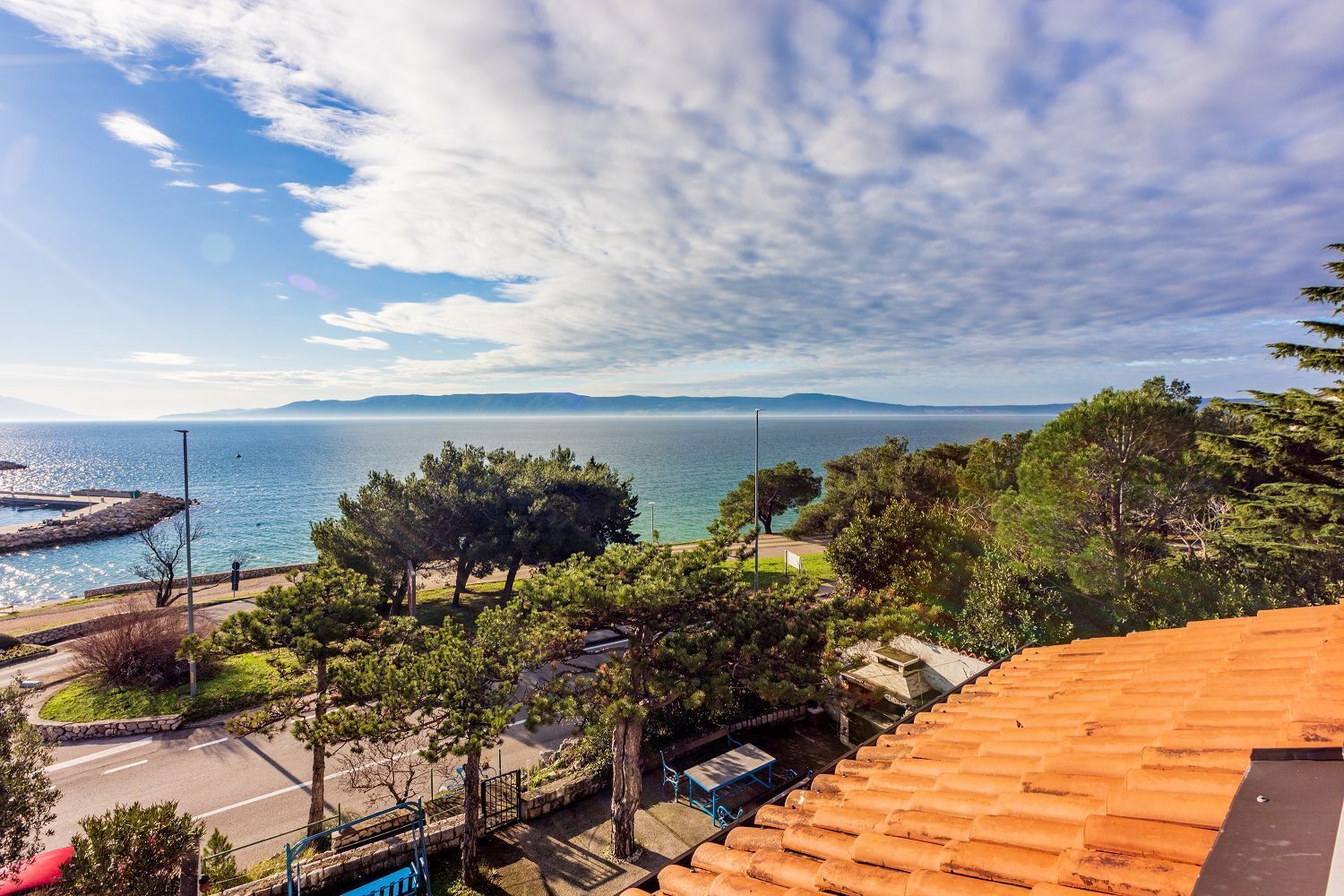 A coastal view with a paved area, trees, and a stone wall.