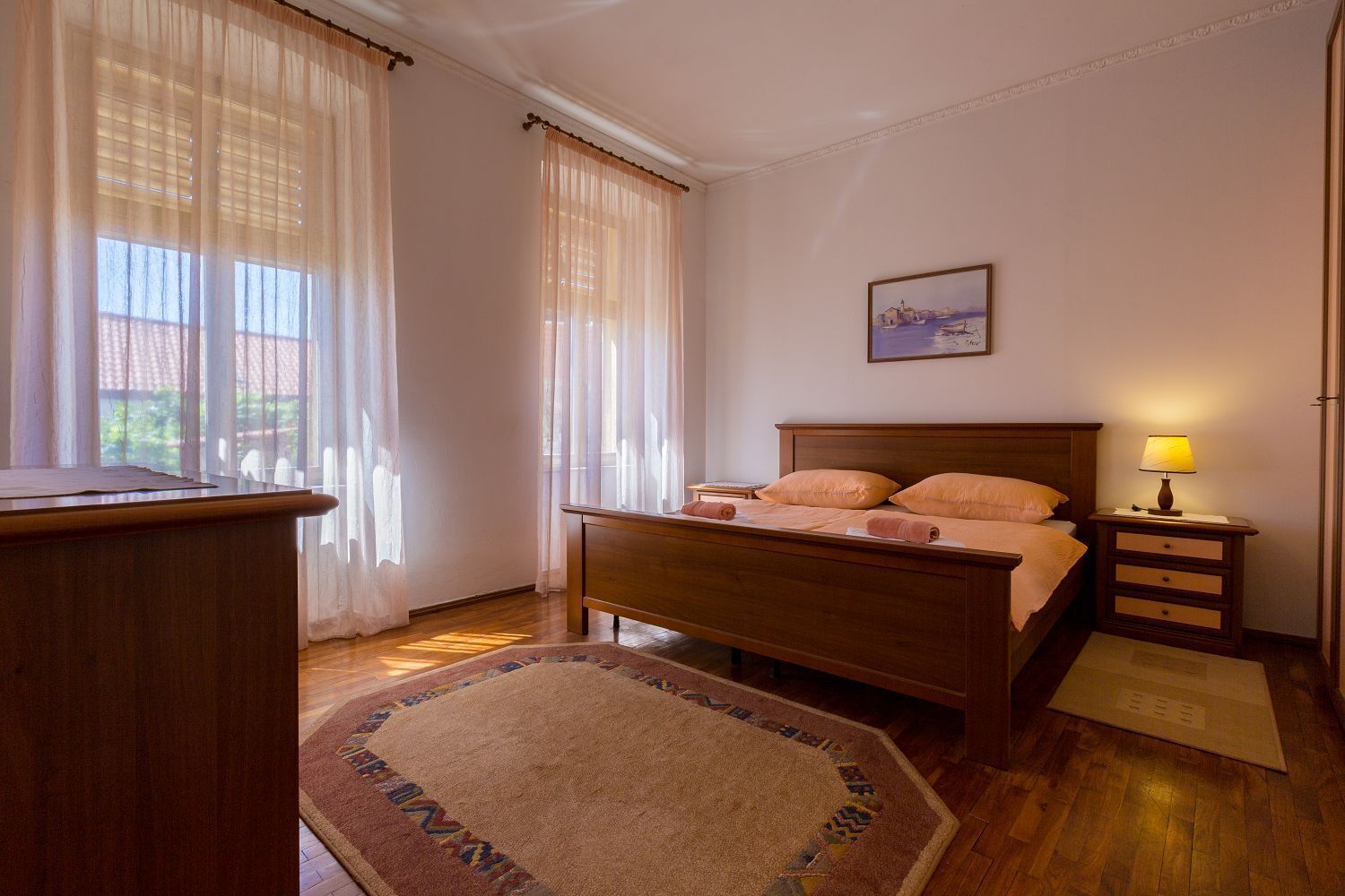 Un dormitorio con cama de madera, una mesita de noche con lámpara, un cuadro en la pared y una ventana con cortinas translúcidas.